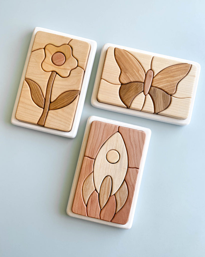 Three Handmade Wooden Puzzles are shown on a flat surface, each crafted from Cherry wood and handmade in Russia. With designs of a flower, butterfly, and rocket, the puzzles feature natural wood tones enhanced by linseed oil, offering simple yet clean images with distinct outlines for each piece.