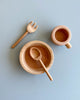 A flat lay of the Handmade Pretend Dinnerware Set with a natural wood finish on a pale blue background, featuring a bowl with a spoon, a cup, and a fork, all arranged neatly and symmetrically.