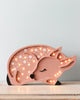 A decorative children's night light shaped like a sleeping fawn is depicted. The Little Lights Deer Lamp is pink with white polka dots and illuminates softly from within, creating a warm and cozy ambiance. Handmade from pinewood, the fawn has its eyes closed, adding to the serene and peaceful effect.