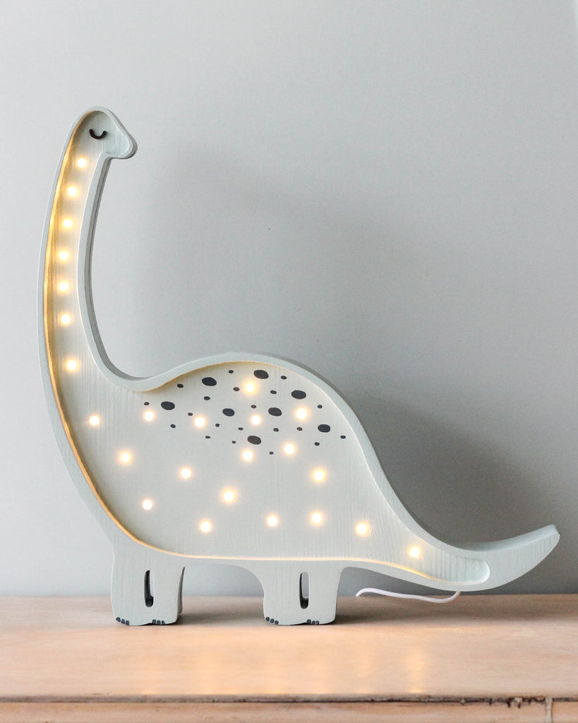 A light blue Little Lights Dinosaur Lamp resembling a brontosaurus stands on a handmade natural pinewood surface. The lamp features illuminated spots along its back and neck, emitting a soft, warm glow—an ideal piece for nursery decor against a neutral background.