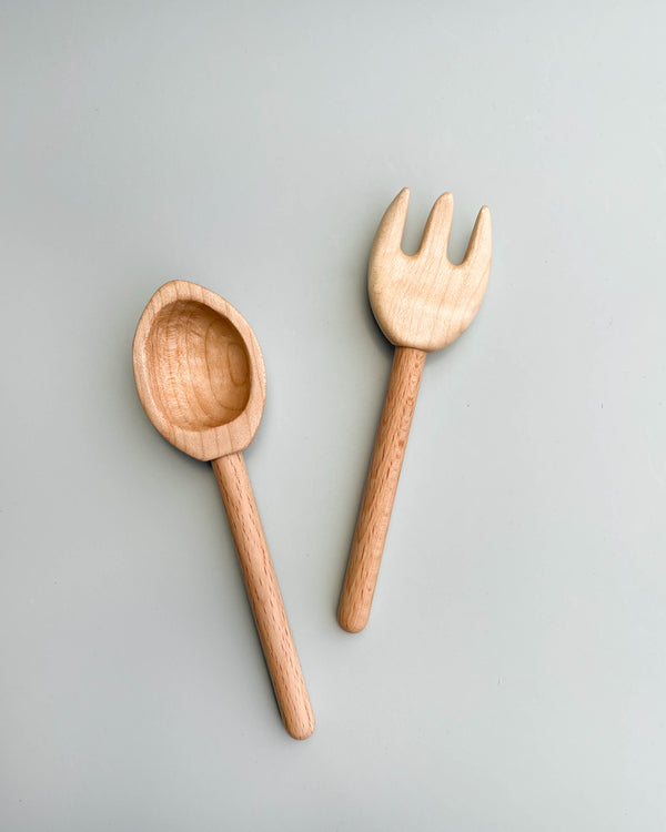 Two handcrafted wooden utensils on a light gray surface: the Handmade Wooden Spoon with a deep, round bowl positioned on the left and the Handmade Wooden Fork with three blunt tines placed on the right. Both feature smooth, cylindrical handles and are made from light-colored wood, perfect for imaginative play.