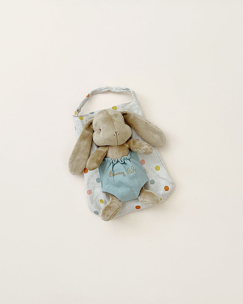 A plush gray Maileg Bunny Bob with long ears, wearing a light blue dress embroidered with "Bunny Girl," is resting inside a matching polka-dotted fabric bag with a handle. The bag and plush toy are placed against a plain, light background.