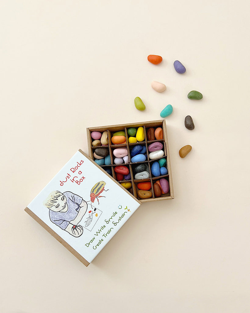 An open box of colorful Non-Toxic 64 Piece Crayon Rocks with a doodled book cover saying "just derps. dreaming of food. little moments of us." Some crayons are scattered beside the box on a