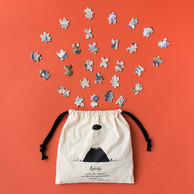 A beige drawstring bag adorned with a black illustration of a volcano and the text "Let's be honest, Ask your grandparents" sits atop an orange background. Above it, scattered pieces from the Londji - Dino Explorer Puzzle (350 pcs) form an arc.