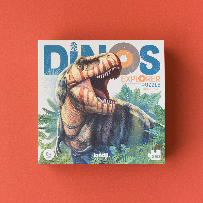 Product photo features a Londji - Dino Explorer Puzzle (350 pcs) against a red backdrop. The box is adorned with an illustration of a roaring dinosaur amid lush green foliage. It includes the text "DINOS EXPLORER Puzzle" and "Look & Learn." This educational toy is designed for children aged 8 and up.