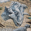 A close-up of a partially assembled Londji - Dino Explorer Puzzle (350 pcs) depicting a detailed illustration of a blue and grey dinosaur labeled "Kosmoceratops." Another smaller dinosaur, labeled "Psittacosaurus," is partially visible in the corner of the image, showcasing an educational toy that sparks curiosity about prehistoric life.