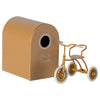 A small brown Maileg Tricycle With Basket with white wheels and a wooden seat is placed next to a small brown cardboard playhouse. The playhouse has a rounded roof and a circular opening on one side with the words "Abri à Tricycle" written on it.