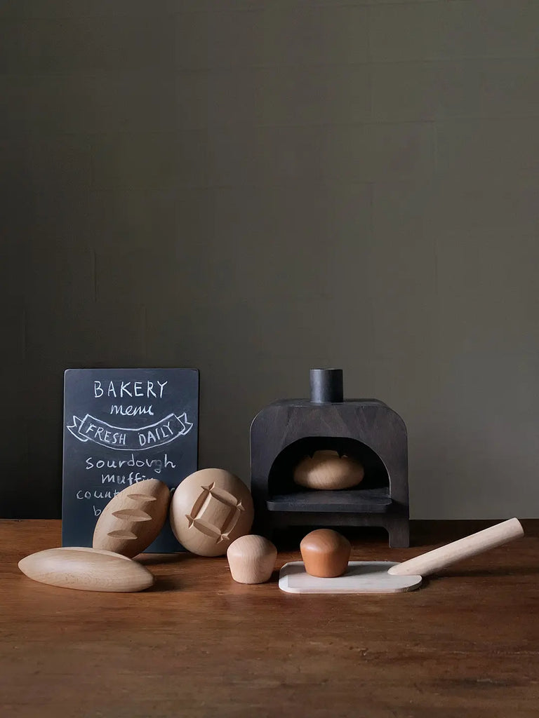 A small tabletop scene featuring My Bakery, a miniature cast iron oven with a loaf of sourdough bread inside, wooden baking utensils, and a raw egg on a wooden surface