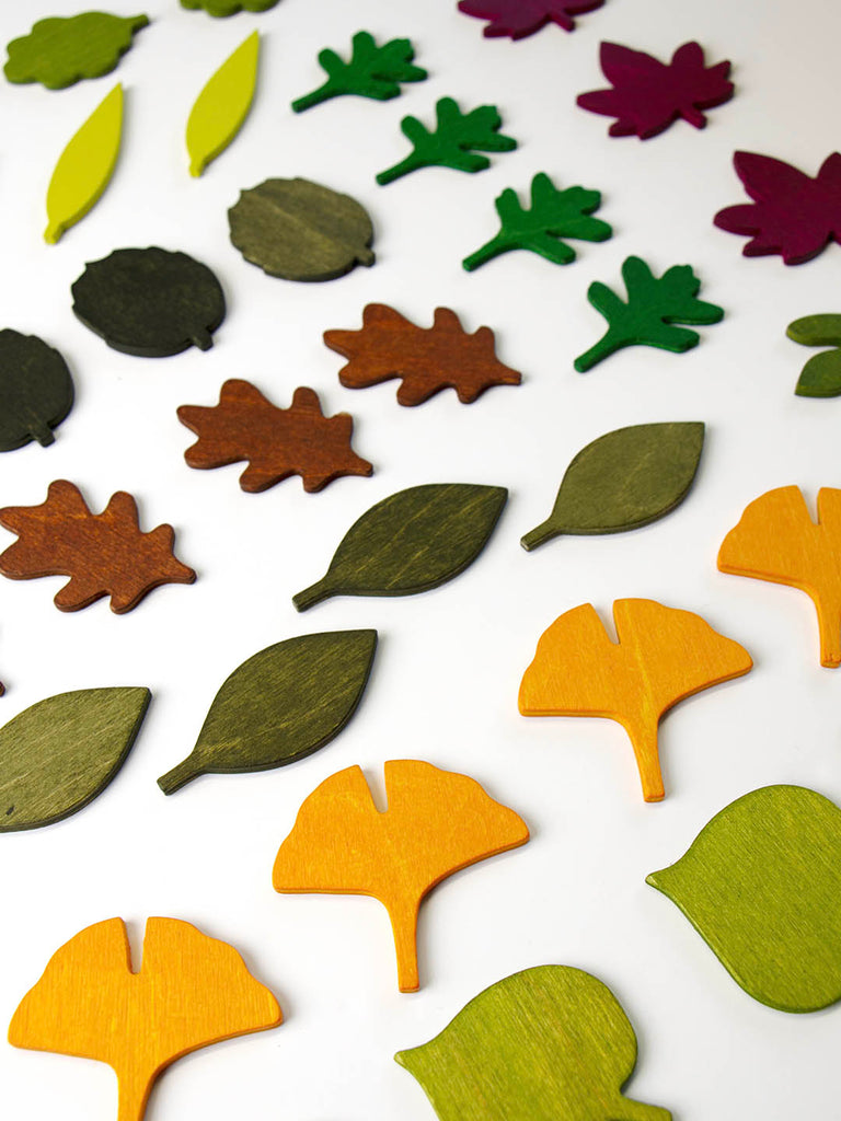 A variety of colorful, wooden leaves are scattered on a white surface. These Woodland Leaves come in different shapes and are painted in shades of green, brown, yellow, orange, and purple, resembling different species and seasons—perfect for loose parts play.