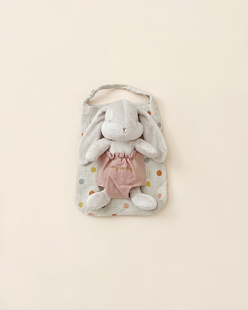 A Maileg Bunny Holly peeking out of a polka dot fabric bag with a pastel pattern on a pale yellow background. The bunny is wearing a pink dress.