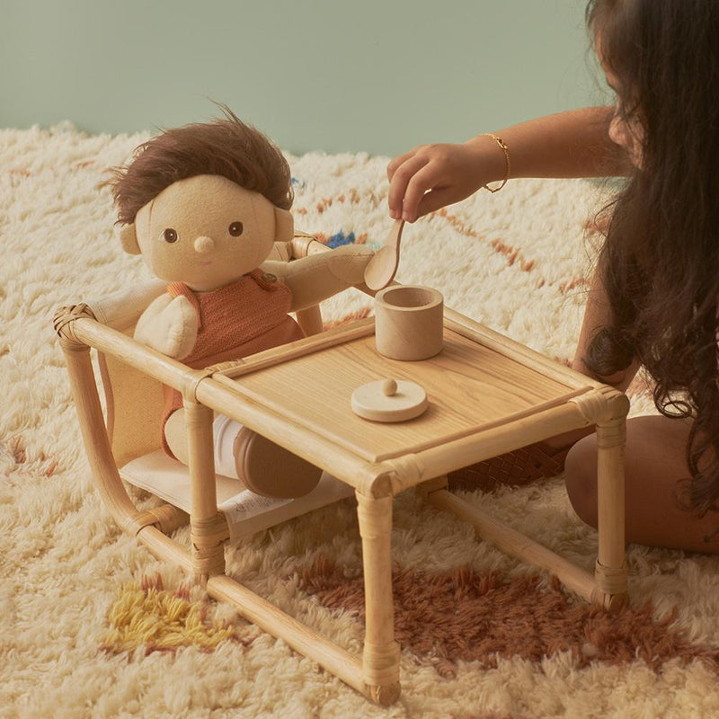 A child plays with a Wooden Doll Feeding Set seated at a small pinewood table, pretending to pour tea from a tiny pot into a cup, on a plush cream-colored rug against a pale green background.