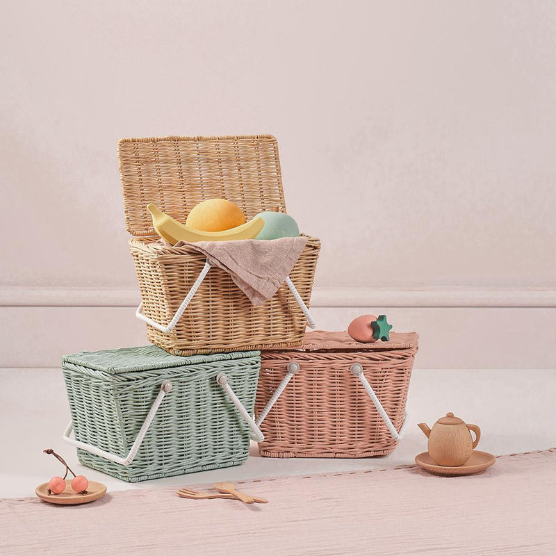 Three colorful hand-woven Olli Ella | Piki Baskets, one stacked open with fruit inside, beside two ceramic teacups and saucers, set against a soft pink background.