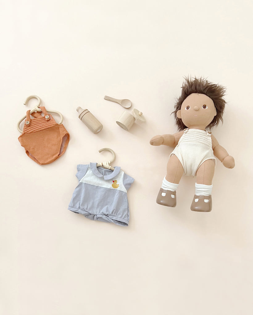 A Olli Ella | Dinkum Doll with dark hair and accessories including a small orange bag, bottle, pair of shoes, and blue cotton pyjamas with a chick design, neatly arranged on a light background.