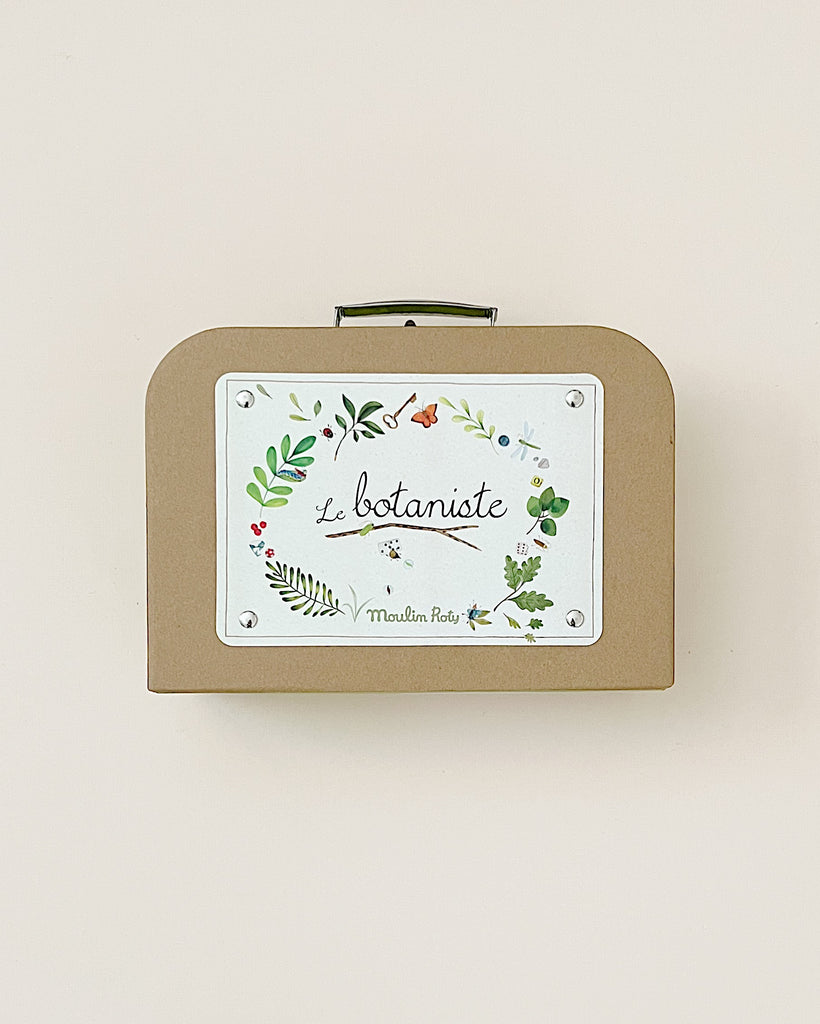 A vintage-inspired Moulin Roty The Botanist Suitcase from the young botanist's garden collection, featuring the words "le botaniste" and floral illustrations on a cream background, set against a plain beige wall.