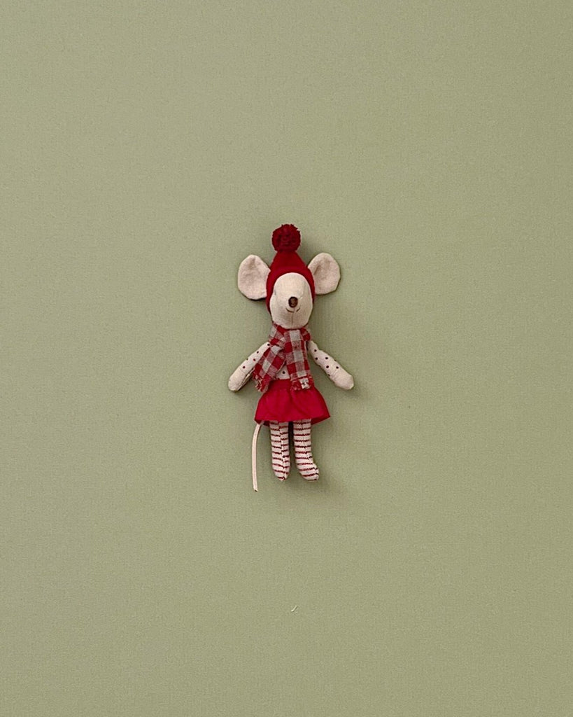 A small Christmas mouse with cream-colored fur is dressed in a red dress and striped stockings. The Maileg Christmas Mouse - Big Sister, your cozy small friend, has large round ears and wears a red hat with a pom-pom. It’s set against a plain light green background, ready to bring festive cheer to any gingerbread house display.