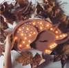 A hand holds the Little Lights Deer Lamp, a children's night light crafted from handmade pinewood, shaped like a sleeping deer with illuminated spots. The glowing light is surrounded by scattered brown maple leaves on a white surface, creating an autumnal atmosphere.