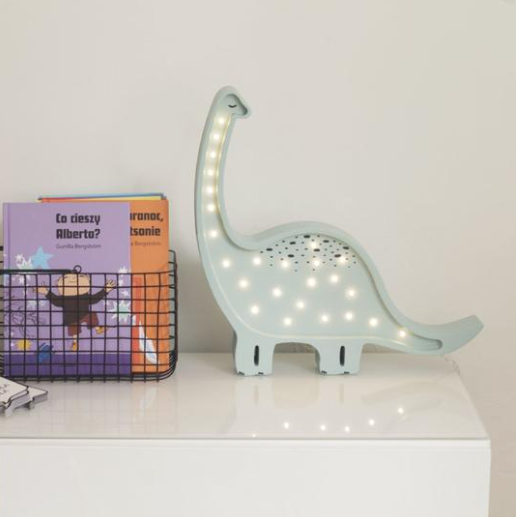 A white table holds a small wire basket filled with children's books. Next to the basket, a light blue Little Lights Dinosaur Lamp made of handmade natural pinewood glows softly. The background is a plain white wall, creating a charming nursery decor.