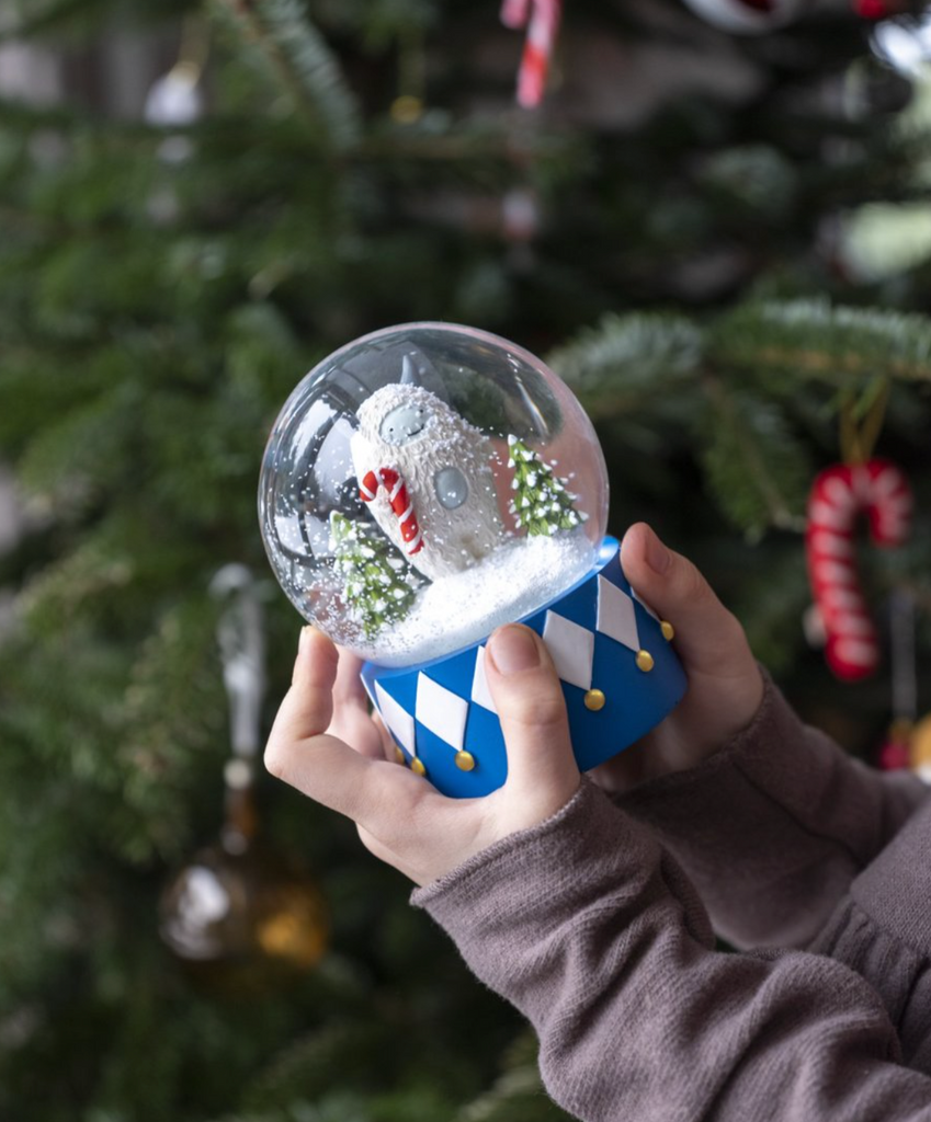 A child holds the Yeti Snow Globe, showcasing a small polar bear and Christmas trees inside. The globe is set on a blue and white checkered base, evoking a magical yeti landscape. In the background, there's a decorated Christmas tree adorned with candy cane ornaments.