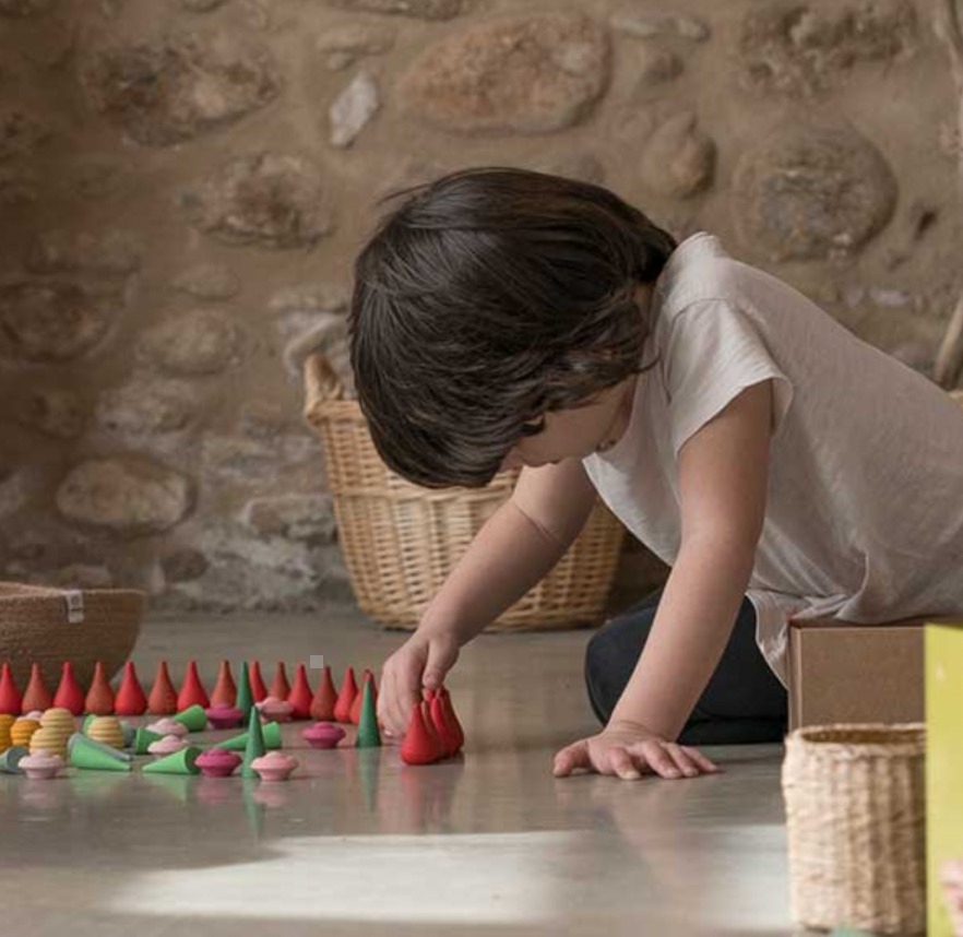 A young child crouches on a stone floor, carefully placing colorful Grapat Mandala Cones Fire, crafted with non-toxic dyes, in a row, with focus and curiosity evident in their expression.