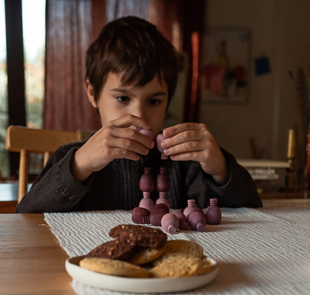 A young boy at a dining table intently focuses on assembling small purple pieces from a Grapat Mandala Pineapples collection, with pretend food on a plate in front of him. The room is dimly lit, creating