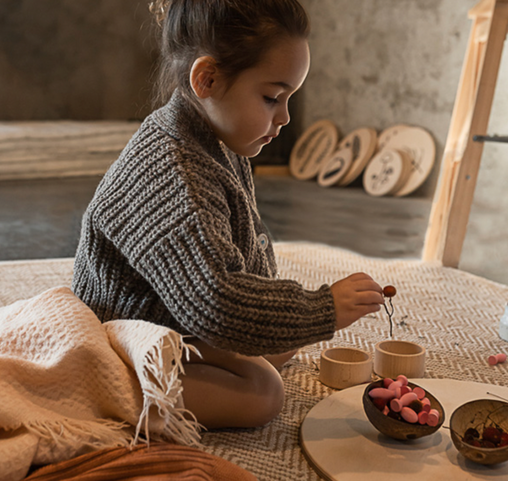 A young girl in a cozy knitted sweater plays with Grapat Mandala Petals on a rustic floor, surrounded by soft blankets and a serene, softly lit atmosphere.