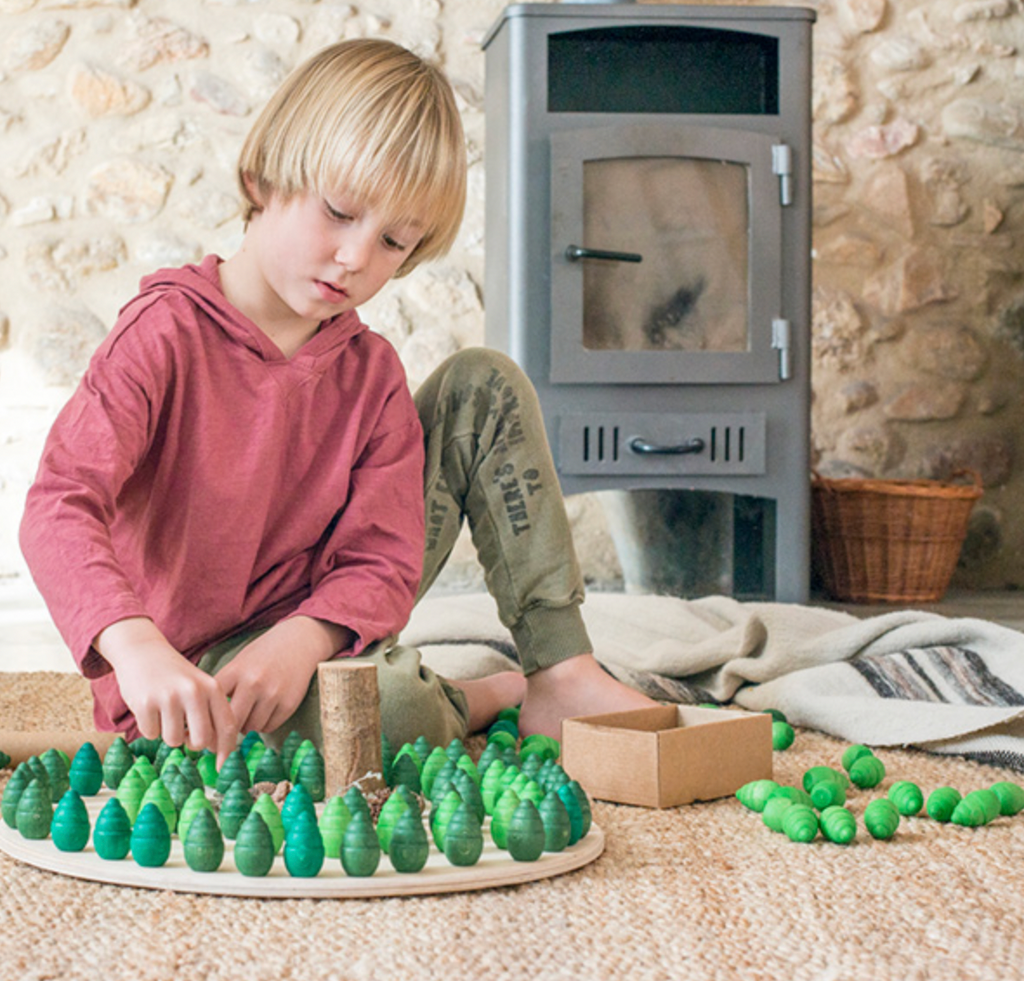 A young child in a red shirt and green pants, surrounded by a blanket and stone walls, plays with Grapat Mandala Trees made from sustainable forests on the floor near a wood stove.