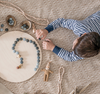 A child in a striped shirt sits on a textured rug, arranging Grapat Mandala Stones and beads into patterns on a circular wooden board made from wood sourced from sustainable forests, surrounded by crafting materials.