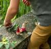 A child in textured pants and a striped shirt engages in imaginative play with the small Grapat Yay! Play Set toys resembling gnome-like figures wearing pointy hats on a moss-covered stone outdoors, one figure adorned with a miniature sn