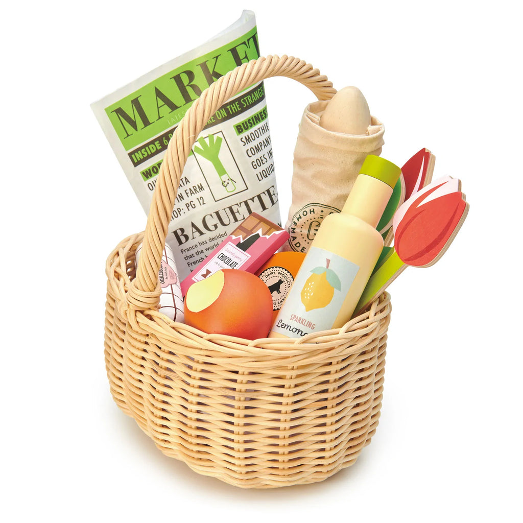 A handcrafted wicker basket filled with toy groceries, including a carton labeled "MARKET," a pretend baguette in a cloth bag, a toy vinegar bottle, a wooden tomato, a lemon drink bottle, and colorful toy fruits and vegetables. Perfect for pretend play, the Shopping Basket (ships in approximately one week) has a handle for easy carrying.