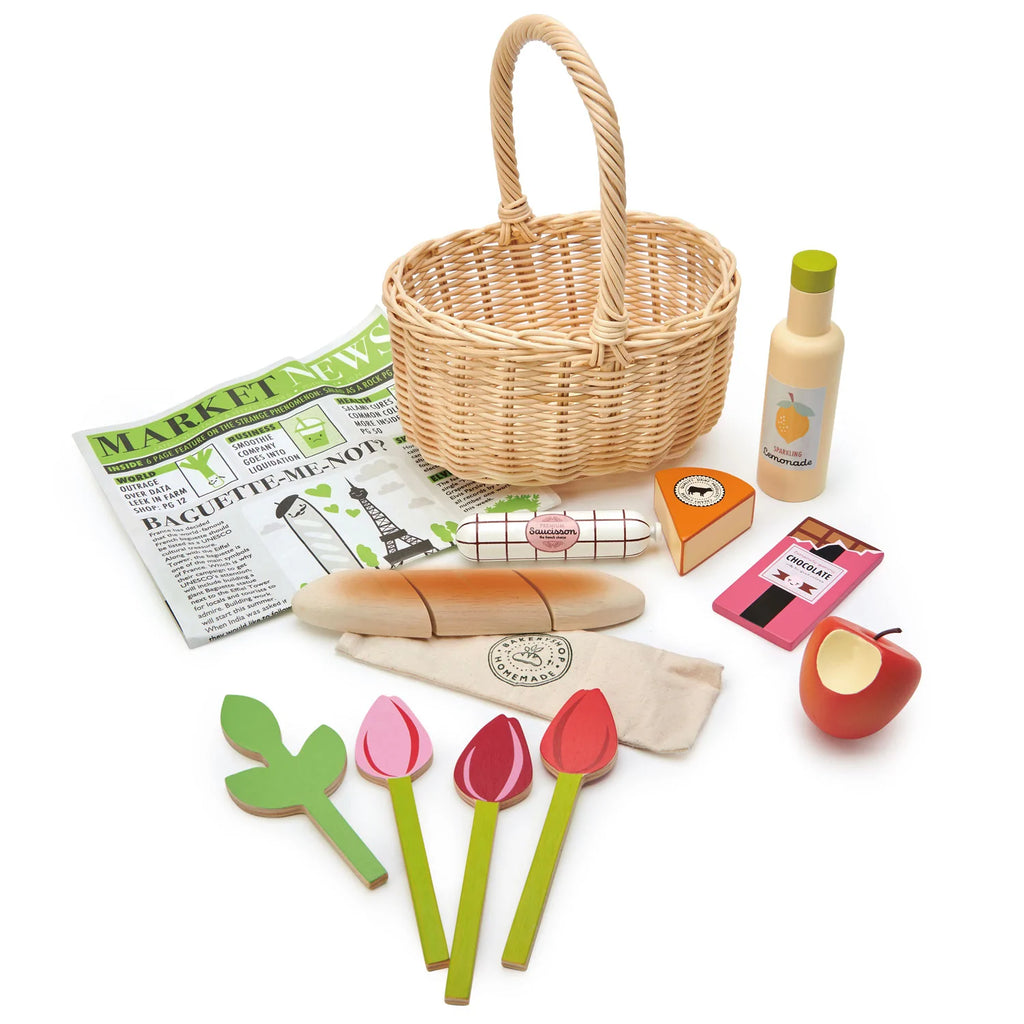 A handcrafted wicker Shopping Basket (ships in approximately one week) surrounded by wooden toy items including three tulip flowers, a green leaf, an apple, a block of cheese, and more - perfect pretend play accessories for children. It also includes a bag with baguette slices, a whole baguette, and a pretend newspaper titled "Market News.