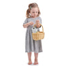 A young girl with light brown hair stands barefoot in a gray dress. She holds a Shopping Basket (ships in approximately one week) filled with vegetables in one hand while examining the contents with the other. The background is white, making her look like she's engaged in delightful pretend play.