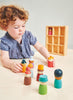 A young child with curly hair and a blue shirt is engaged in open-ended play with colorful Happy Folk Hotel figurines on a table. Behind the child is a solid wood shelf with multiple compartments, one of which has a single Happy Folk Hotel figurine inside it.