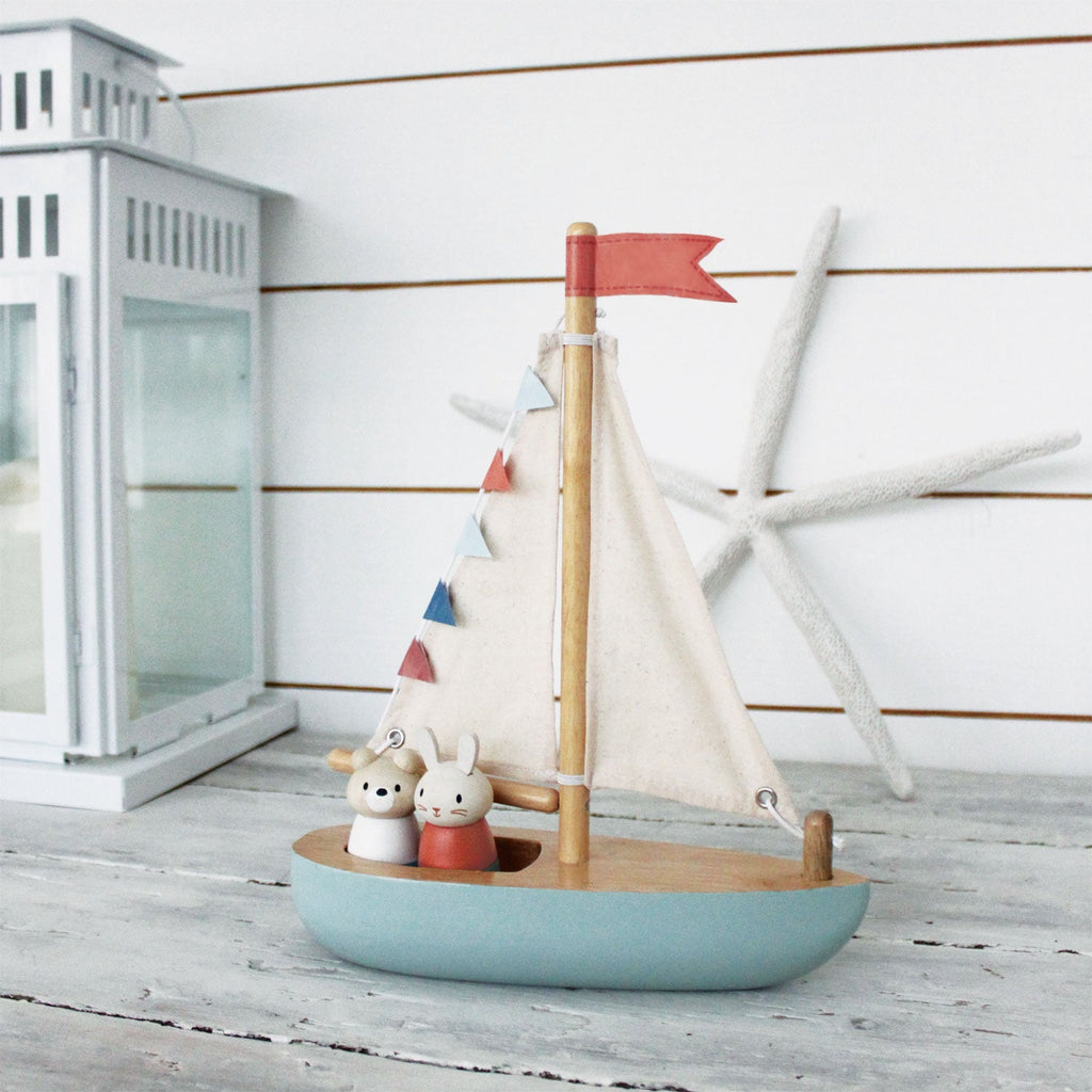 A Wooden Sail Boat with a blue hull, fabric sails marked with red and blue colors, and two small figures on board. The boat is displayed in front of a white paneled wall.