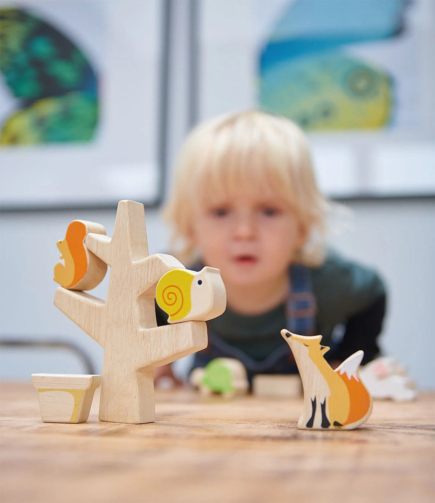 A young child with blonde hair, blurred in the background, intently observes Stacking Garden Friends arranged on a table, featuring a solid wood tree-shaped toy holder in focus.