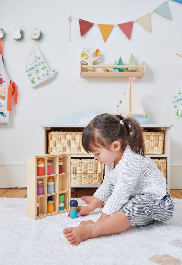 A young girl with pigtails is sitting on a white rug engaging in creative play with the Happy Folk Hotel. Behind her, there is a shelf with baskets and various decorative items on the wall, including a string of colorful flags, toys, and hanging bags designed to inspire open-ended play.