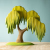 A photo of a stylized, handmade Large Wooden Willow Tree with a brown trunk and drooping, layered green foliage. The children's tree toy is set against a light blue background and stands on a wooden surface. The design is simple and artistic, featuring non-toxic paint with exaggerated leaf shapes.