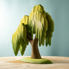 A smooth wooden sculpture of a willow tree, designed as a children's tree toy, featuring a textured trunk and leafy branches cascading down. The Large Wooden Willow Tree is placed on a wooden surface and set against a light blue background, finished with non-toxic paint for safe play.