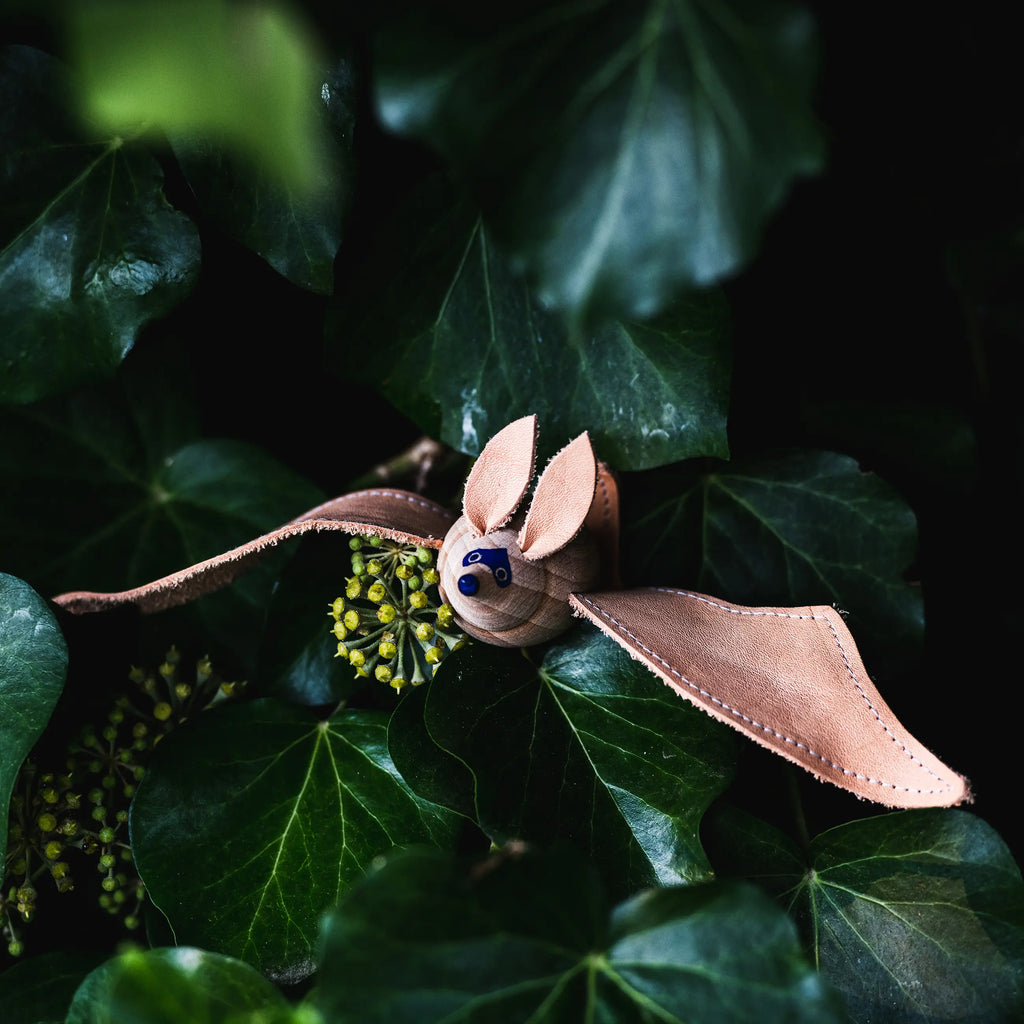 A Handmade Wooden Bat, crafted from light brown leather with elongated leather wings, is placed amidst dense green foliage. The bat has pointed ears and dark button-like eyes, giving it a cute and whimsical appearance.