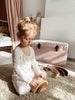 A young child with light brown hair tied up in a bun sits on a carpeted floor, engaged in open-ended play with the Handmade Rainbow Stacker - Pink. She is wearing a white long-sleeve shirt and white pants adorned with small pink dots. In the background, there is a pink trunk and a bed draped with a pink curtain.