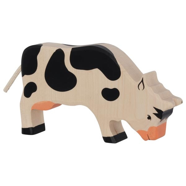 A Holztiger Grazing Black Cow with a natural wood finish and painted black and orange patches. Crafted in Europe, this toy features a simple, stylized design with a tail made of rope. Like other HOLZTIGER figures, it lacks intricate details, giving it a simplistic, rustic appearance.