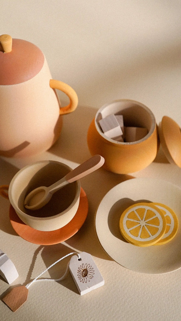 A cozy still life scene with a Handmade Wooden Tea Set - Flower including a teapot, cups, and plates in earthy tones, adorned with lemon slices and a geometric tag, bathed in soft sunlight