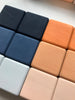 A close-up of nine square-shaped, wooden blocks arranged in a grid. The blocks, painted with non-toxic paint, vary in color—including dark blue, light blue, navy, peach, light peach, and beige. These Mini Wooden Blocks - Desert Night have a smooth texture and are placed on a white surface.