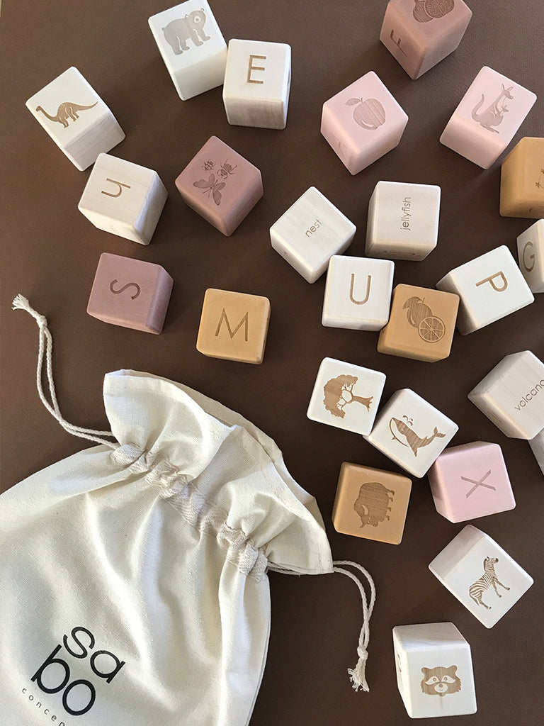 An assortment of Alphabet Wooden Blocks - Rose with letters and animal engravings scattered around a small canvas drawstring bag on a brown surface.