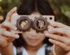 A person holds a Twinkle Kaleidoscopic Camera up to their face. The background is out of focus, showing greenery. The person's fingernails are painted, and their hair is black. Crafted from high-quality walnut and beech wood, the image conveys a playful and creative atmosphere.