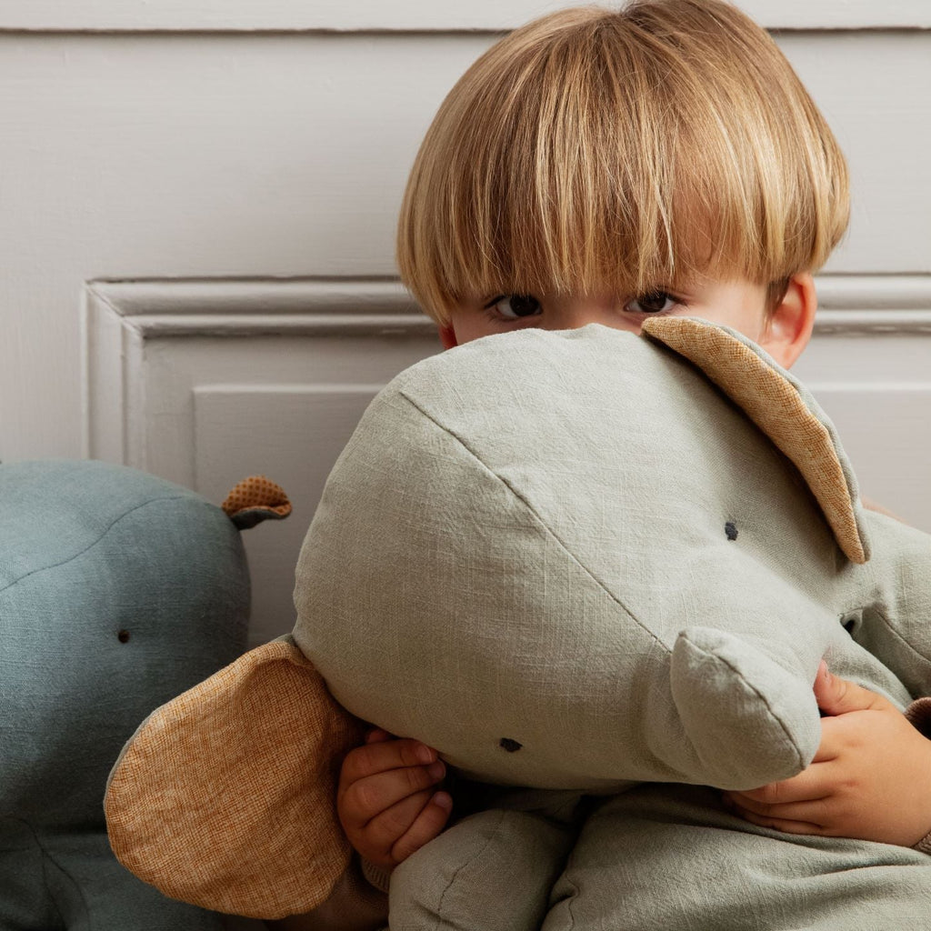 A young child with blond hair, peeking over a Maileg Big Grey Elephant toy, eyes visible above the elephant's head, with a soft focus background.