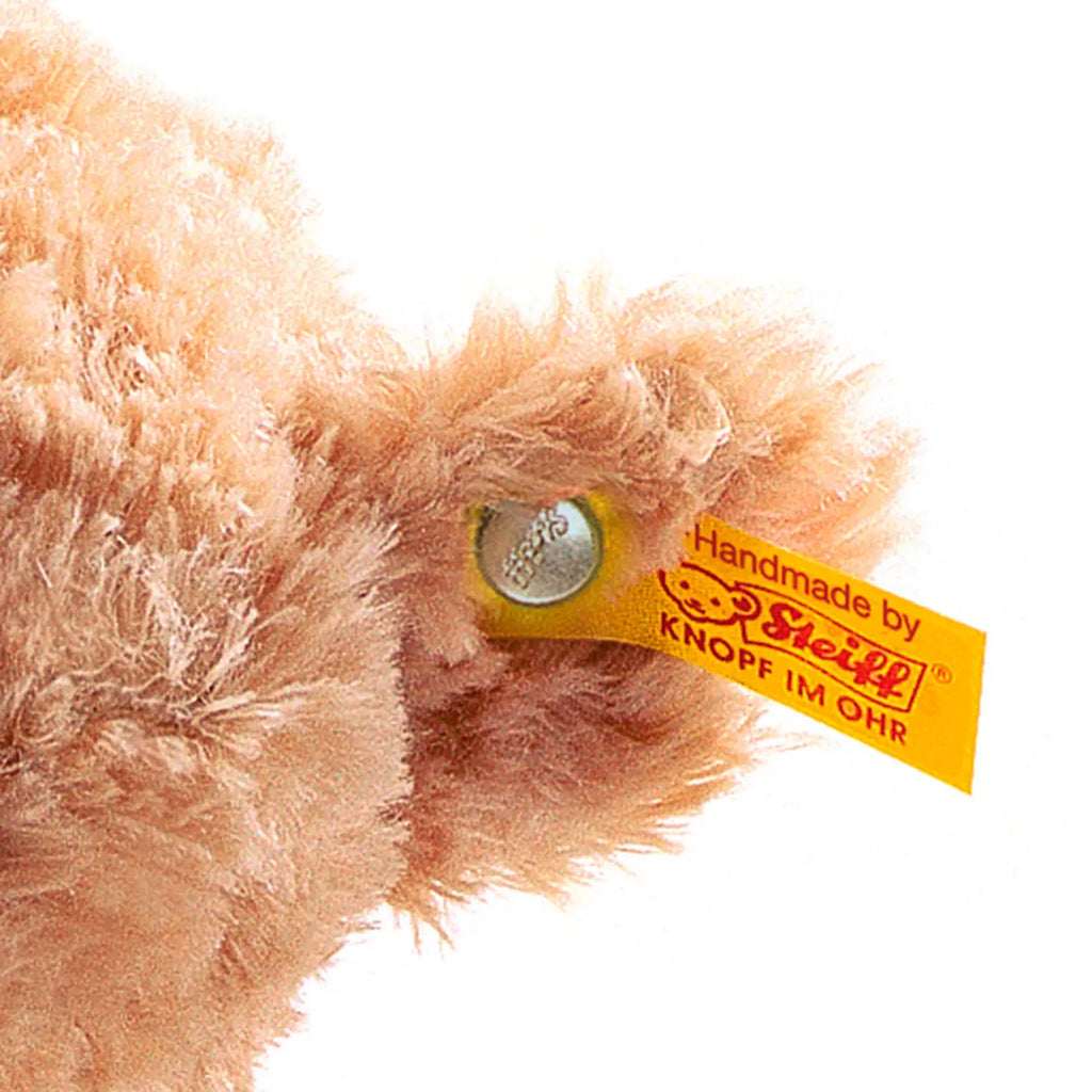 Close-up of Elmar Teddy Bear's ear showing the brand's trademark yellow tag and "Button in Ear," indicating authenticity. Elmar Teddy Bear's fur is soft and light brown.