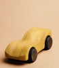 A Handmade Wooden Race Car - Yellow crafted from Beech with a smooth, natural finish and dark wooden wheels sits on a plain beige background. The car has sleek, simple lines and a natural wood grain pattern accentuated by non-toxic paint.