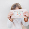 A child holding the Retro Wooden Water Game in front of their face, obscuring it. The frame displays an abstract, colorful painting. The background is a plain, light-colored wall.