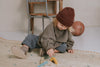 A young child wearing a red beanie and a grey sweater sits on a patterned rug indoors, playing with a Wooden Safari Set, fostering fine motor skills development. A wooden chair and an old basketball are in the background.