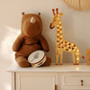 A plush rhino toy sits next to the Little Lights Giraffe Lamp on a white dresser. A small tambourine featuring a rabbit illustration is also placed on the dresser. Warmly illuminated by the Little Lights Giraffe Lamp, which is crafted from natural pinewood, the scene exudes a cozy atmosphere.